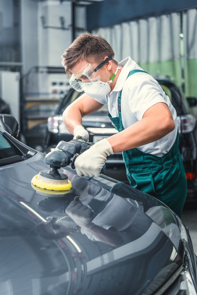 A worker in protective gear polishes a car with a power buffer in a well-lit garage, focusing on a glossy surface.