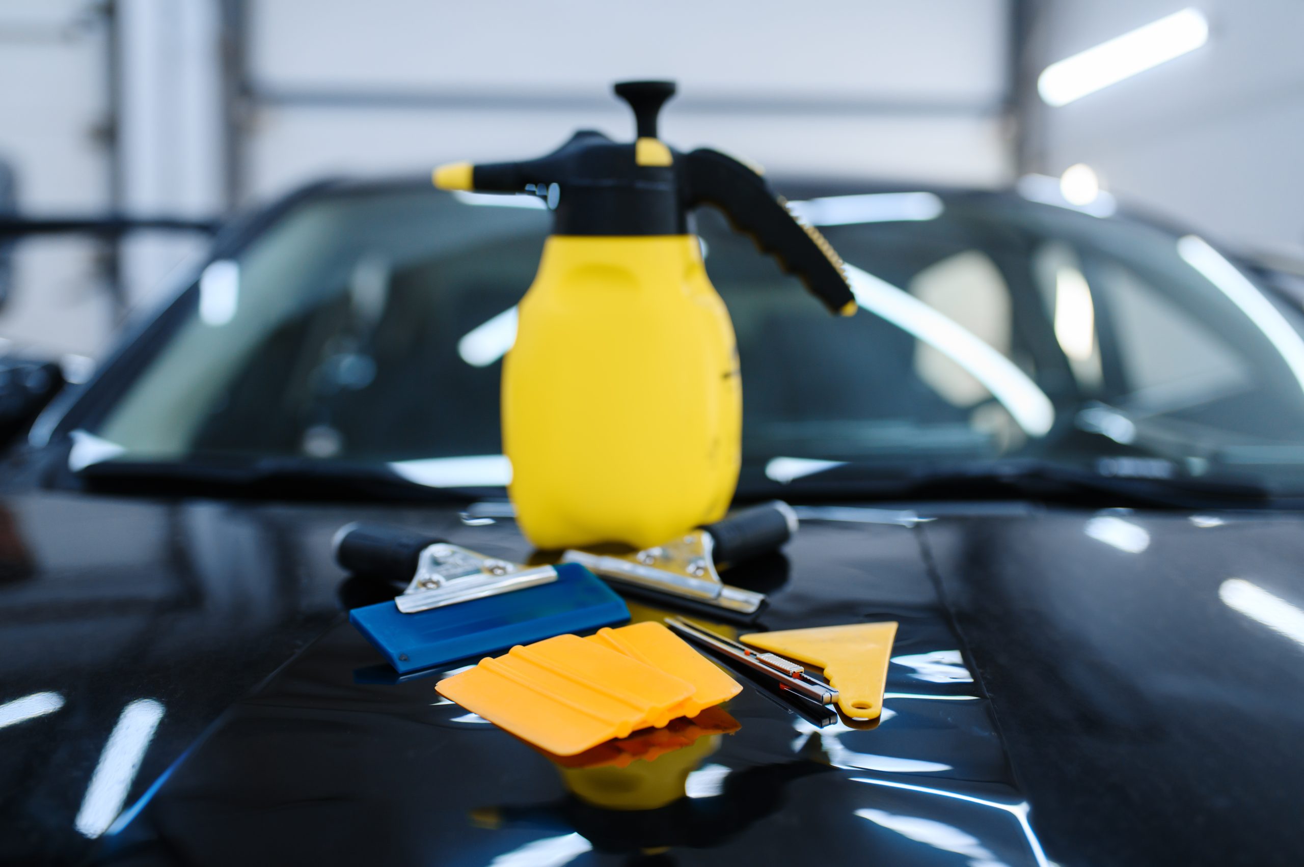 A yellow spray bottle sits on a black car hood, surrounded by various squeegees and scrapers in blue and orange hues.