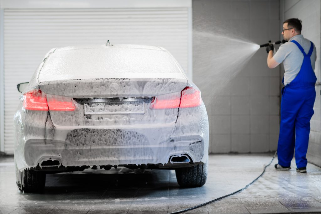 A man in blue overalls washes a foam-covered car from behind in a clean garage, spraying it with a pressure washer.
