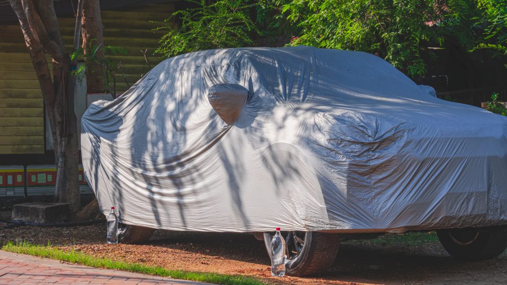 A car covered with a protective cover under a tree, shielding it from direct sunlight to prevent paint fading.